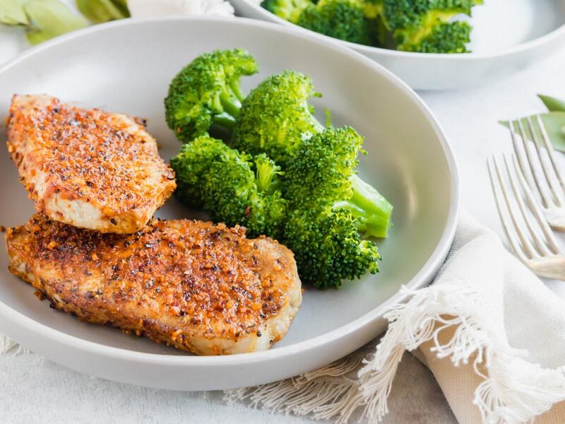 Two cooked boneless pork chops on a dinner plate next to steamed broccoli.