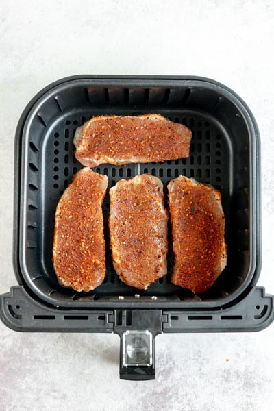 Looking into an air fryer basket with four raw, but seasoned, boneless pork chops inside.