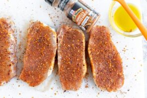 Four pork chops seasoned next to a bowl of oil and a bottle of grill seasoning.