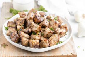 A plate filled with steak bites and topped with parsley sitting on a cutting board.