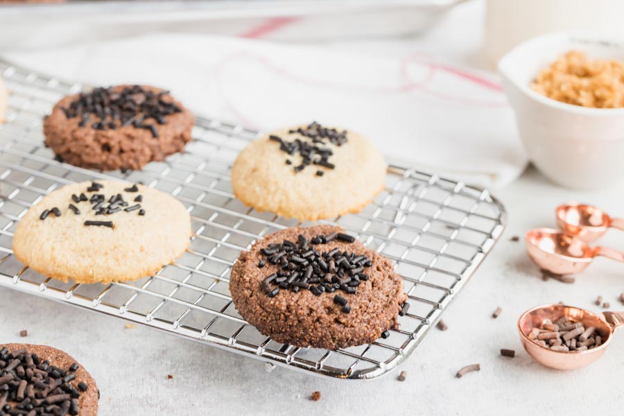 Chocolate and vanilla cookies on a wire rack next to chocolate sprinkles in measuring spoons.