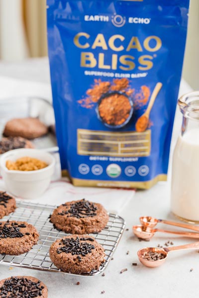 A bag of Cacao Bliss behind a wire rack with chocolate cookies on it. A cup of milk is nearby.