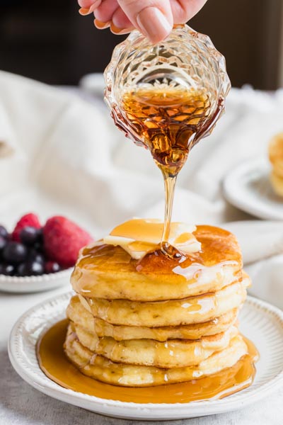 Pouring maple syrup on a stack of pancakes on a plate. Berries are in the background.