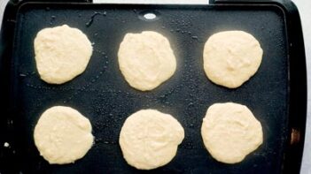 Six pancakes cooking on a non-stick griddle.
