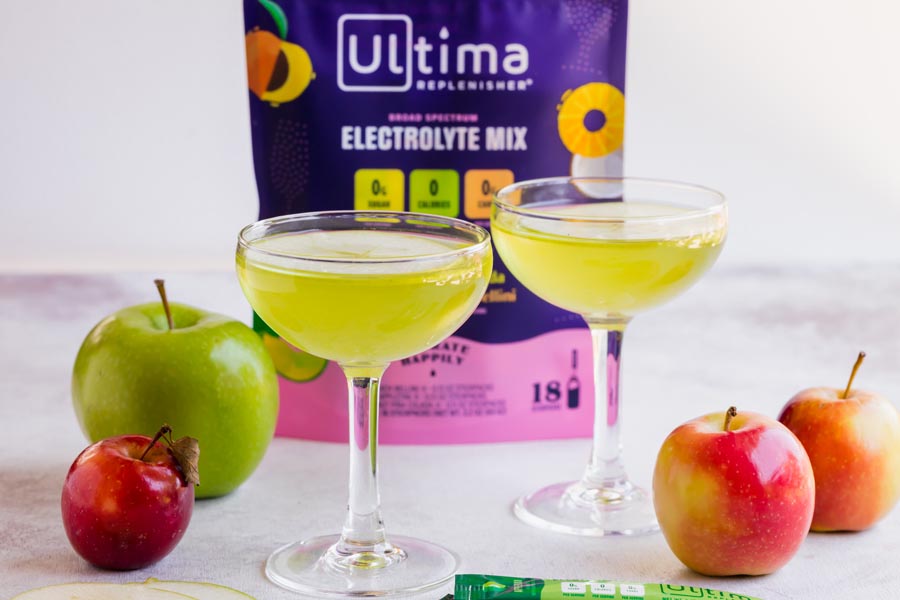 two green cocktails with apples around and a bag of Ultima electrolyte mix in the background