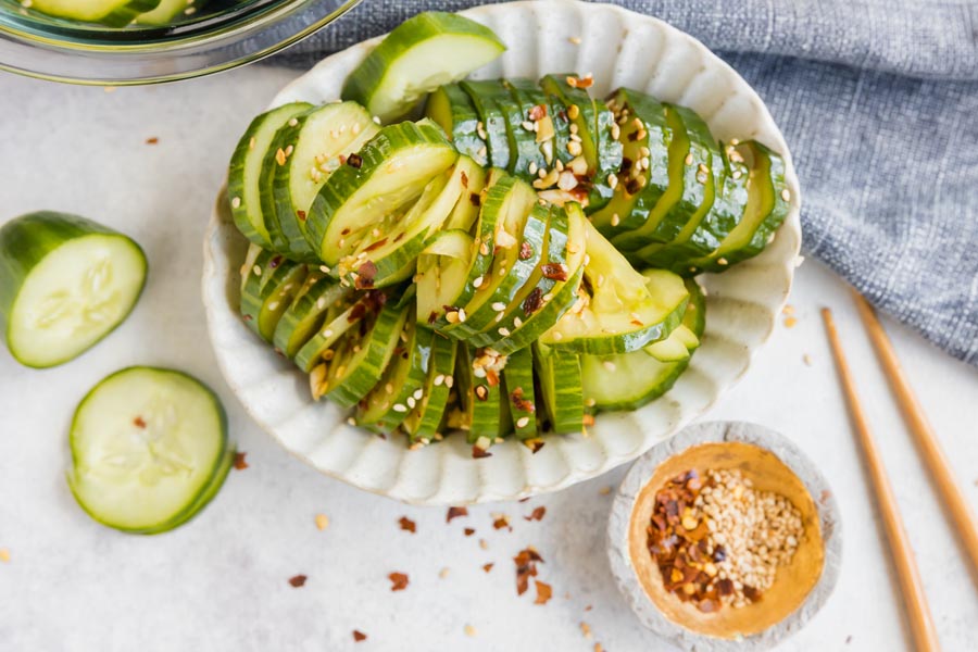 a bowl of accordion cut cucumbers topped with sesames and red pepper next to a bowl of those and sliced cucumbers