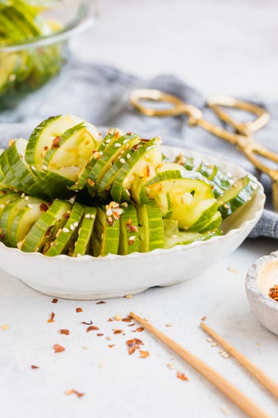 a bowl of sliced cucumbers next to red pepper flakes and chopsticks