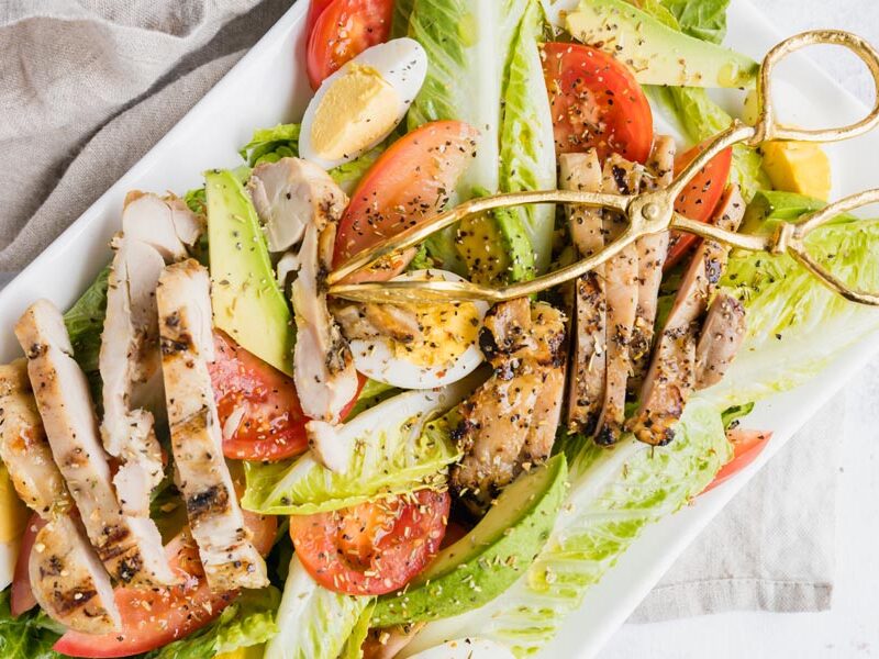 grilled chicken, hard boiled egg, tomato and avocado side on a bed of lettuce with gold tongs on top