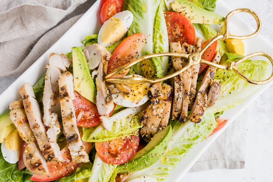 grilled chicken, hard boiled egg, tomato and avocado side on a bed of lettuce with gold tongs on top
