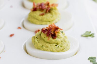 Close up of an avocado filled deviled egg, topped with crumbled bacon and pieces of cilantro near by.