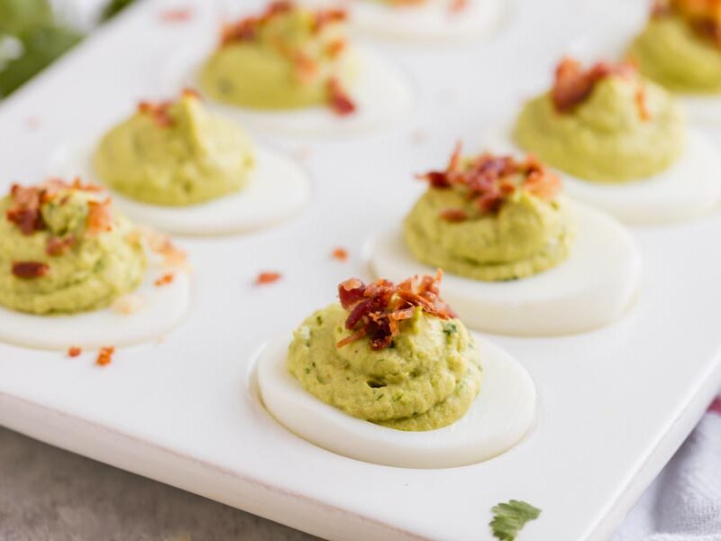 Avocado deviled eggs on a platter with chunks of crumbled bacon on top and sprinkled around.