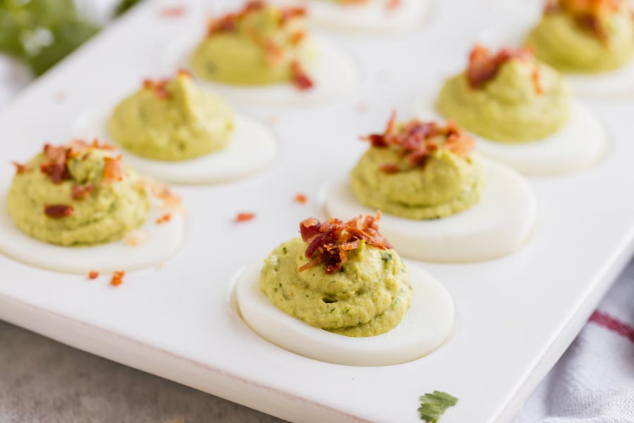 Avocado deviled eggs on a platter with chunks of crumbled bacon on top and sprinkled around.