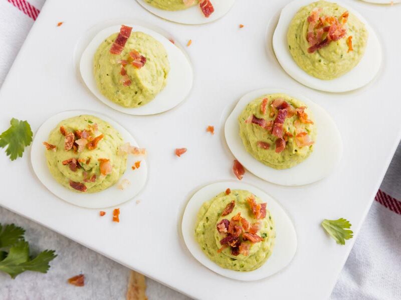 Looking down onto six deviled eggs filled with a green avocado filling and topped with bacon with pieces of cilantro nearby.