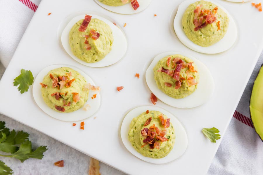 Looking down onto six deviled eggs filled with a green avocado filling and topped with bacon with pieces of cilantro nearby.