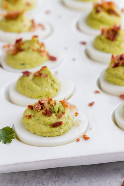 Deviled eggs filled with a green avocado mixture and topped with cooked and crumbled bacon in a deviled egg dish.