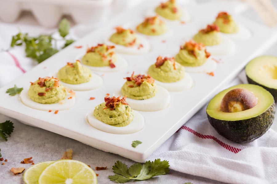 A tray of avocado filled deviled eggs with a half an avocado cut next to the tray and sliced limes.
