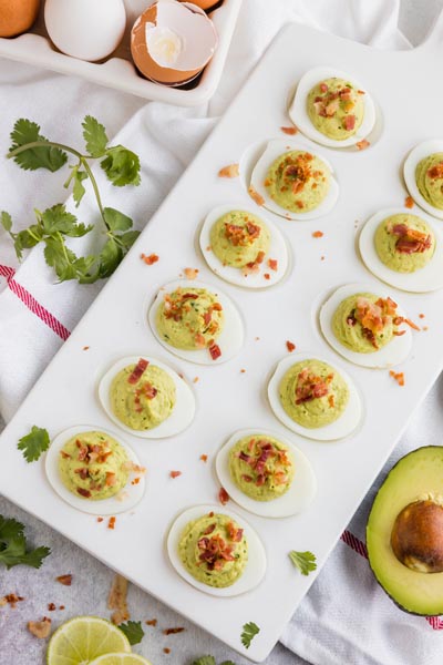 A tray of deviled eggs topped with bacon with avocado, limes and cilantro near by.