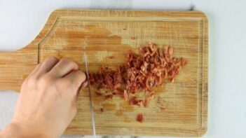 A hand holding bacon while slicing it with a knife on a wooden cutting board.
