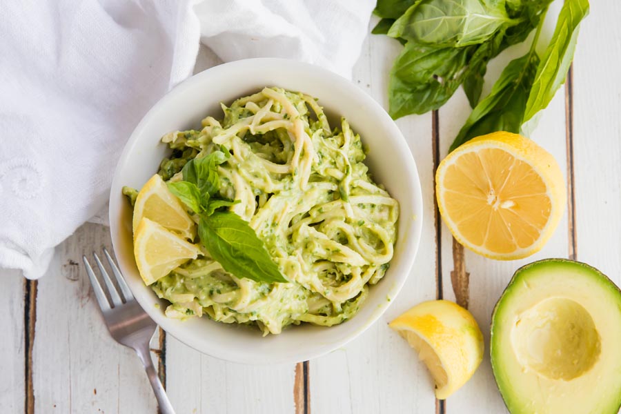 a big bowl of noodles with a creamy green pesto sauce mixed in and lemon garnishes nearby
