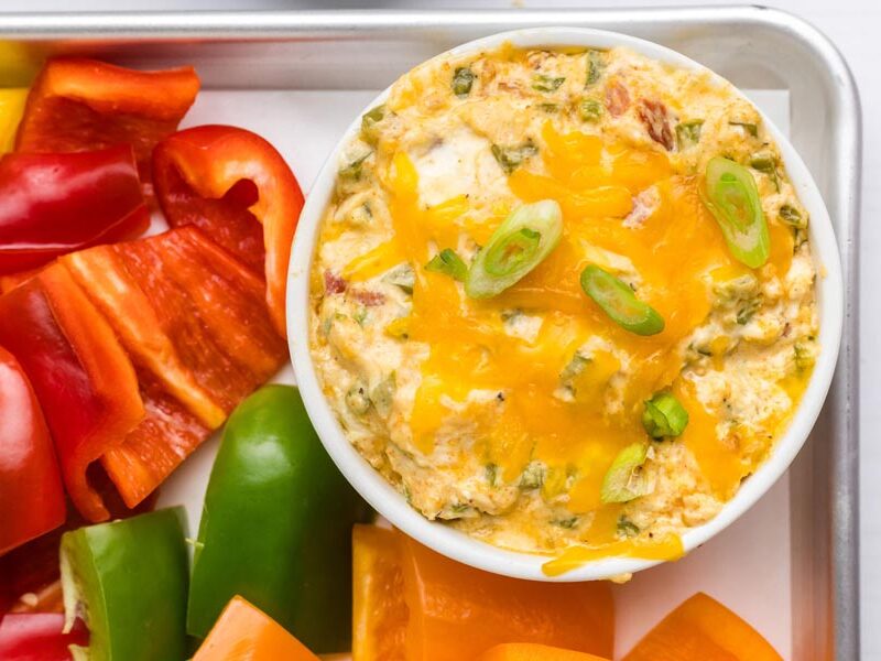 Bacon jalapeño popper dip topped with cheese and green onions beside bell peppers.