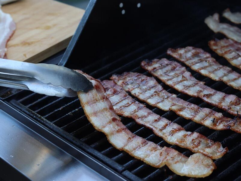 holding a strip of bacon with tongs while strips are cooking on a grill