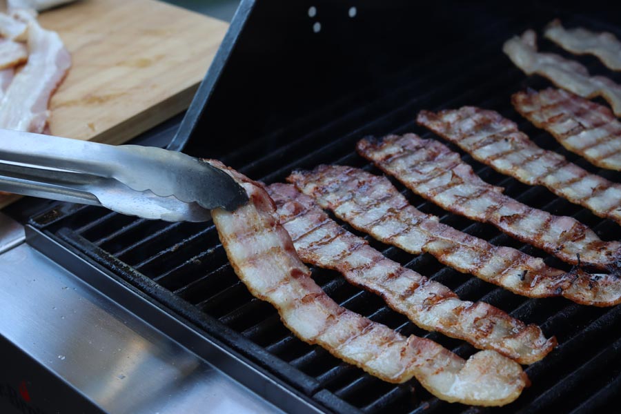 holding a strip of bacon with tongs while strips are cooking on a grill
