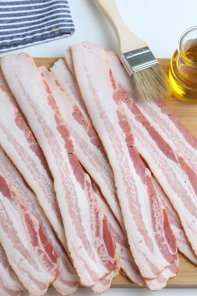 uncooked bacon on a cutting board next to a small jar of oil and a brush