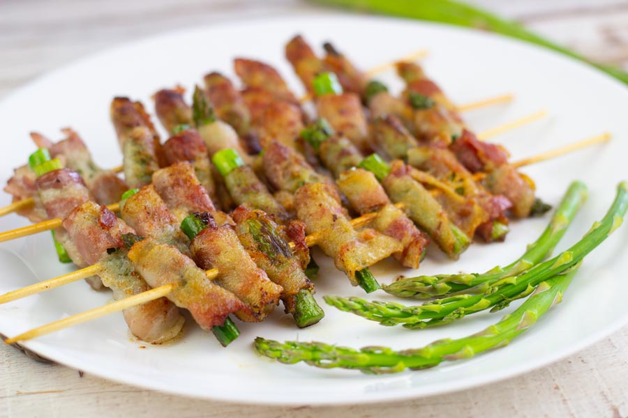 bacon wrapped asparagus skewers on a plate with raw stems of asparagus