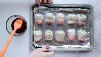 holding a baking tray lined with foil with 10 bacon wrapped brussels sprout on it next to a bowl of maple syrup and an orange brush sticking out