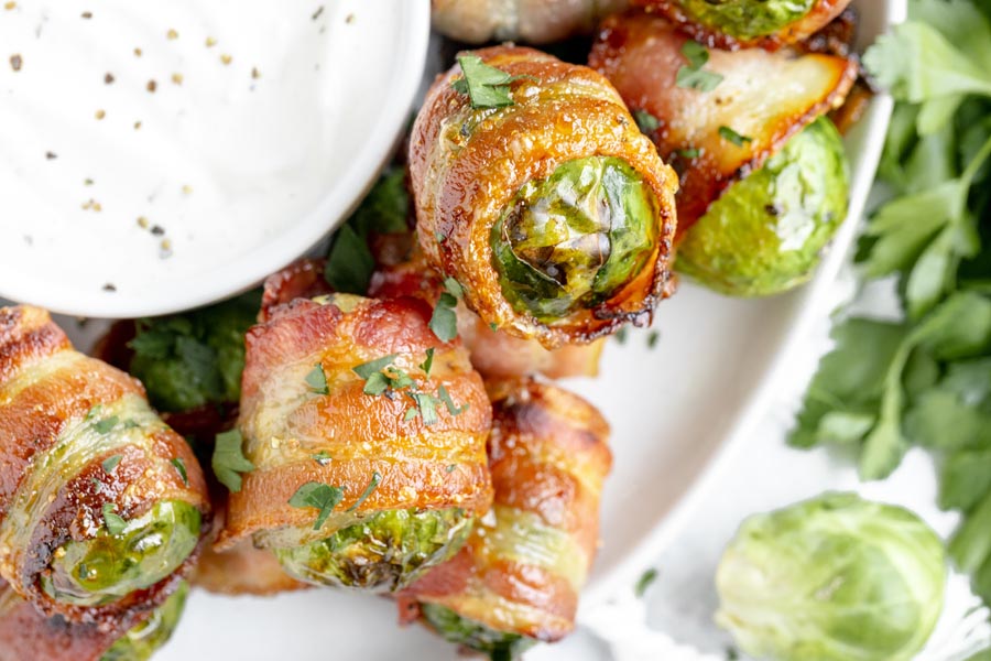 bacon wrapped brussels sprouts it in a serving tray with a bowl of ranch dressing in the center and topped with crumbled feta cheese