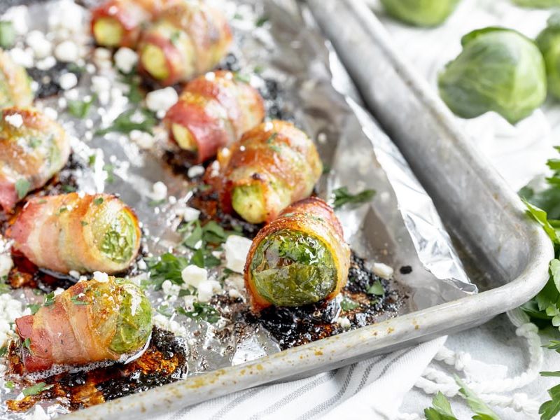 a baking tray with a bunch of bacon wrapped brussels sprouts lined up topped with parsley and feta cheese
