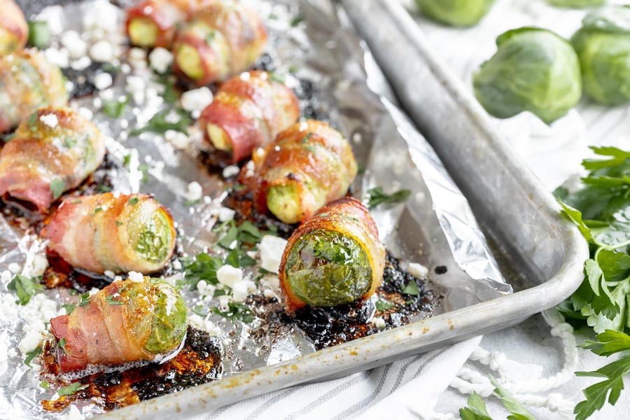 a baking tray with a bunch of bacon wrapped brussels sprouts lined up topped with parsley and feta cheese