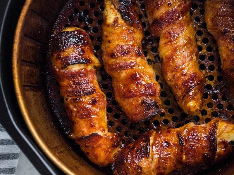 Five bacon wrapped chicken tenders in an air fryer basket. Bacon is crispy and chicken is juicy.