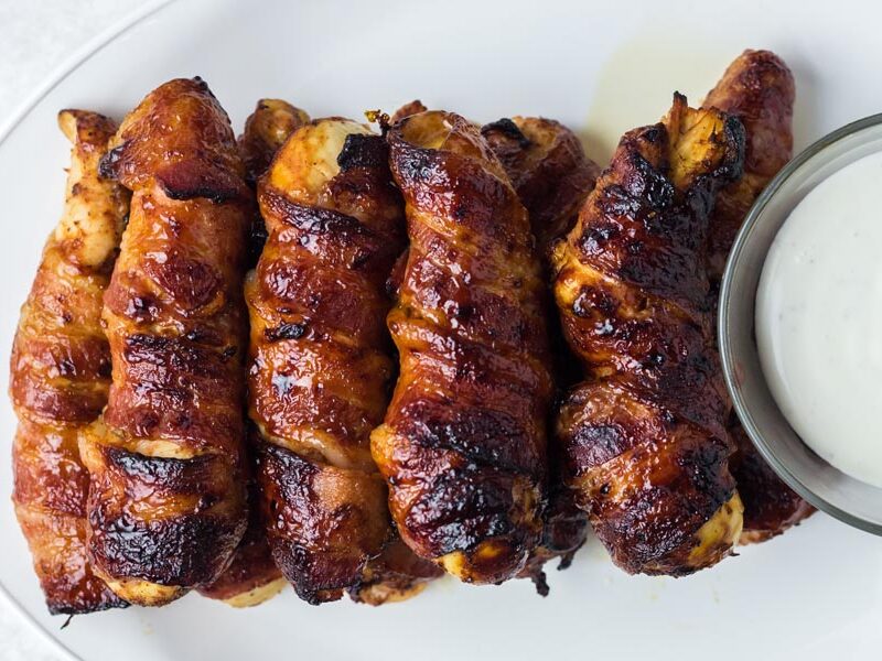 Bacon wrapped chicken stacked on a plate next to a bowl of ranch.