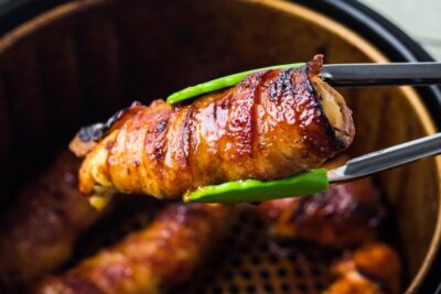 Green tongs holding a chicken tender wrapped in crispy bacon. Juice is dripping from the chicken.