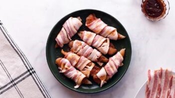 Bacon strips wrapped around pieces of chicken on a plate with raw bacon and bbq sauce nearby.