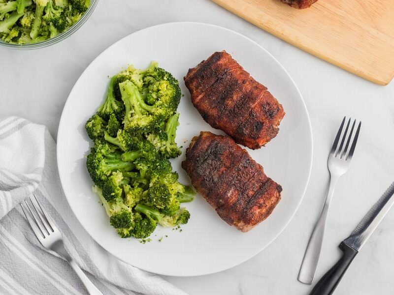 Two bacon-wrapped pork chops with steamed broccoli on a white plate.