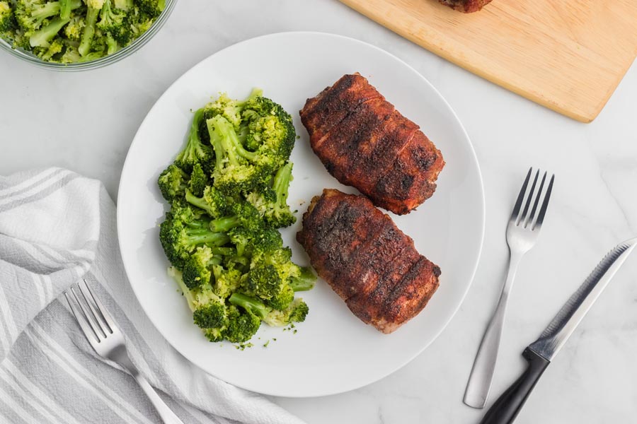 Two roasted bacon wrapped pork chops on a plate with a side of steamed broccoli. Forks and knives are nearby.