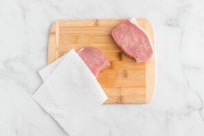 Two pork chops on a cutting pork. One gets patted with a paper towel.