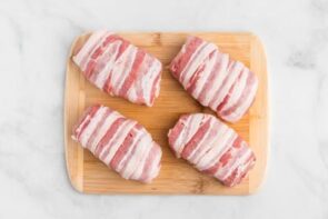 Four pork chops wrapped in bacon on a cutting board.