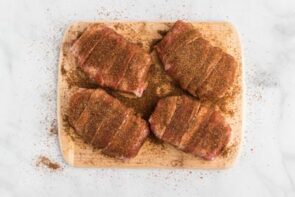 Seasoning rubbed all over the top of bacon wrapped pork chops sitting on a cutting board.