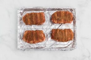A foil lined baking tray holds a wire rack that has four raw bacon wrapped pork chops on top.