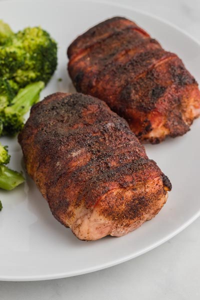 Two boneless pork chops wrapped in crispy bacon with a sweet and spicy seasoning on top next to cooked broccoli.