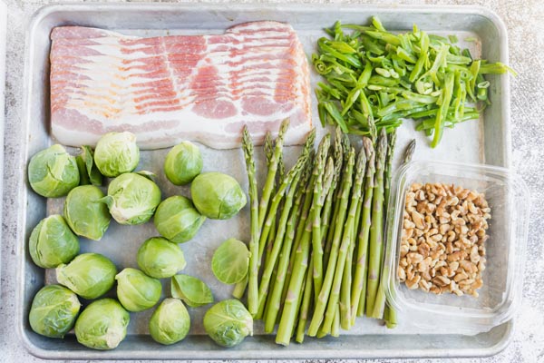 ingredients like asparagus, brussels sprouts, green beans, walnuts and bacon for a sheet pan recipe on a tray