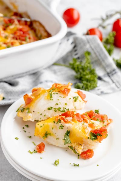 two pieces of baked cod on a stack of white plates topped with tomatoes and parsley