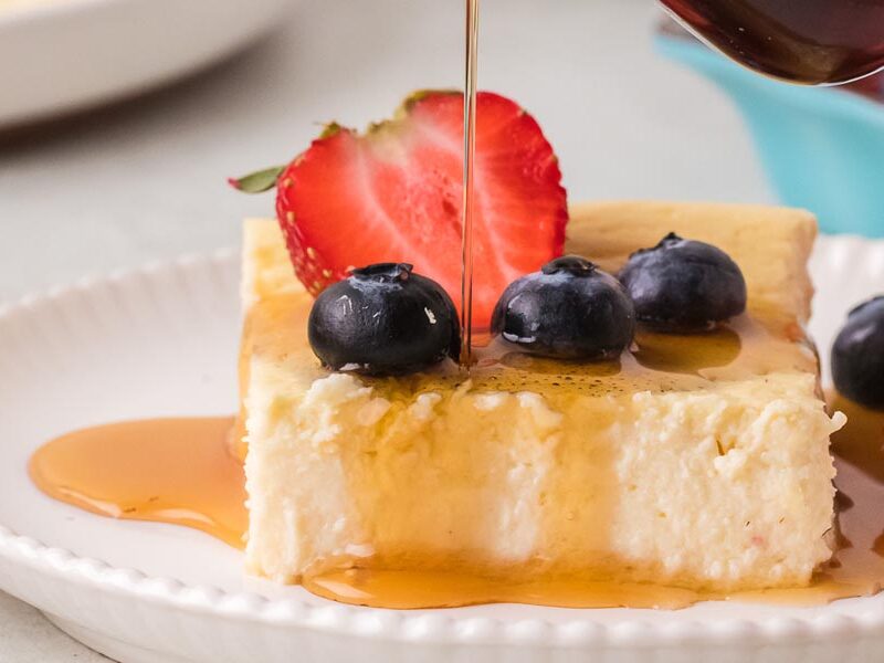 Maple syrup being drizzled over a slice of baked yogurt topped with fresh strawberries and blueberries.