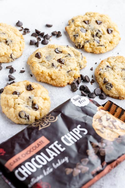 a bag of chocolate chips in front of five chocolate chip cookies with chips spread around