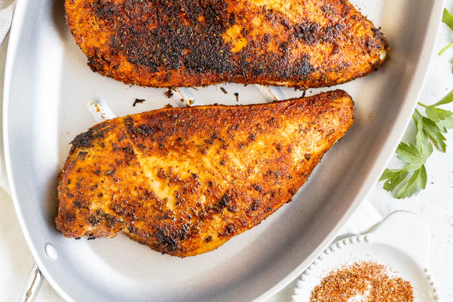 Cooked chicken breasts coated with a charred seasoning of spices on a skillet with a dish of more blackened seasoning nearby.