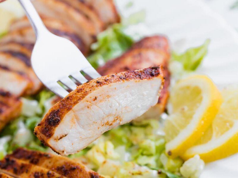 A fork holding a juicy slice of cooked chicken in front of a chicken salad with lemon on the side.