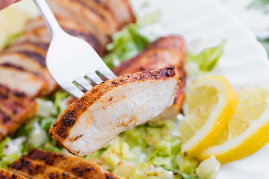 A fork holding a juicy slice of cooked chicken in front of a chicken salad with lemon on the side.